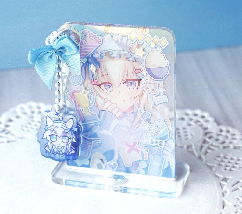 May include: A rectangular acrylic standee featuring a light-haired anime character with purple eyes, decorated with various stickers including a trophy and bowl. A light blue ribbon and a small, dangling acrylic charm are attached to the side.