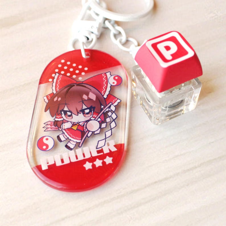 May include: A keychain featuring a red oval acrylic charm with a cartoon character illustration, the text POWER, and three stars. Attached is a white metal ring and a small, clear mechanical keyboard keycap with a red top and the letter P printed in white.