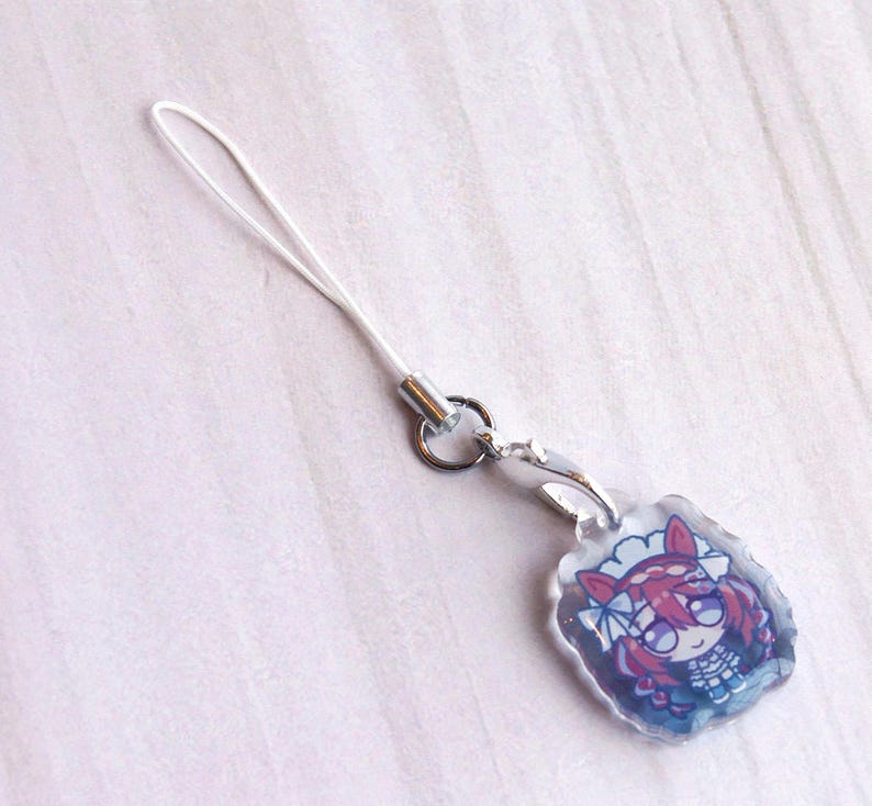 May include: A clear acrylic charm featuring a cartoon character with red hair and animal ears, attached to a silver-toned metal lobster clasp and a white nylon loop cord. The charm has a scalloped edge and a glossy finish over the printed design.
