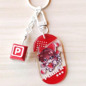 May include: A white metal keychain featuring a red and white oval acrylic charm with an anime-style character illustration, the text POWER, and three stars. A smaller red square charm with a white letter P is attached to the white metal chain.