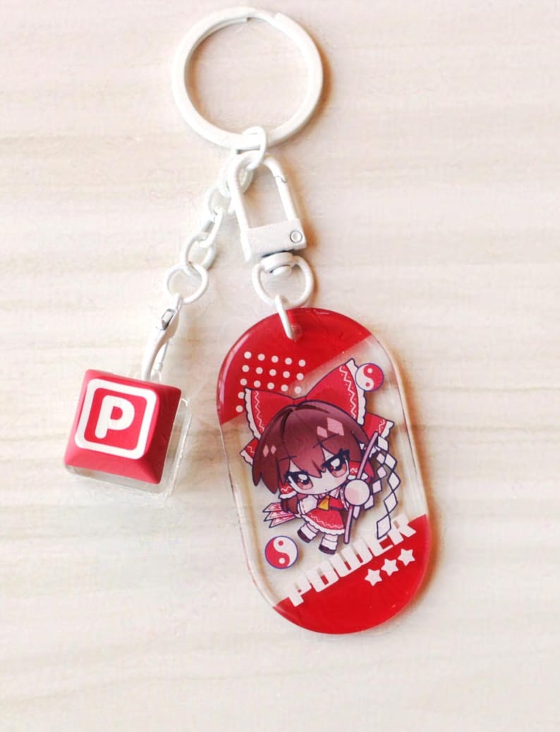 May include: A white metal keychain featuring a red and white oval acrylic charm with an anime-style character illustration, the text POWER, and three stars. A smaller red square charm with a white letter P is attached to the white metal chain.