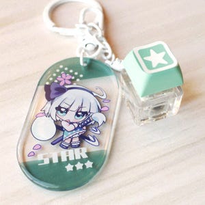 May include: An acrylic keychain featuring an oval charm with a cartoon character illustration, the text STAR, and three stars. Attached is a white metal ring and a light green square mechanical keyboard keycap with a white star icon on top.