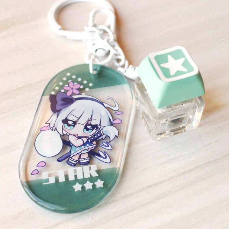 May include: An acrylic keychain featuring an oval charm with a cartoon character illustration, the text STAR, and three stars. Attached is a white metal ring and a light green square mechanical keyboard keycap with a white star icon on top.