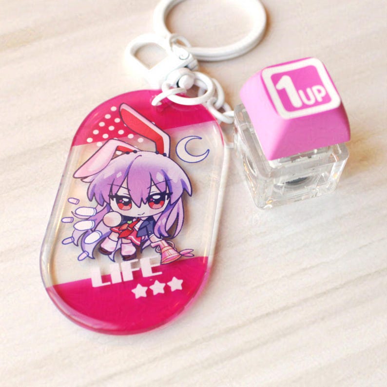 May include: A keychain featuring an oval acrylic charm with a purple-haired character wearing bunny ears, the word LIFE, and three stars. Attached is a clear cube charm topped with a pink square piece featuring the white text 1UP.