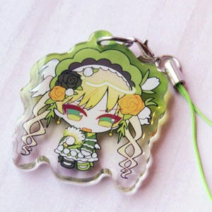 May include: A clear acrylic charm featuring a stylized anime character with blonde hair, green eyes, and a green and white outfit. The character is adorned with black and orange roses and a ruffled headpiece. It includes a silver clasp and a lime green cord.