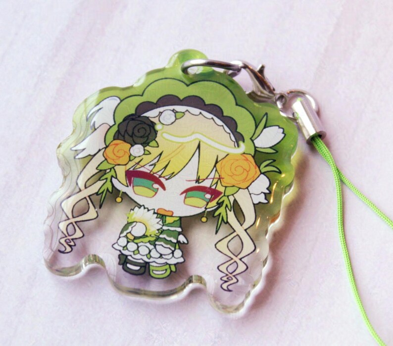 May include: A clear acrylic charm featuring a stylized anime character with blonde hair, green eyes, and a green and white outfit. The character is adorned with black and orange roses and a ruffled headpiece. It includes a silver clasp and a lime green cord.