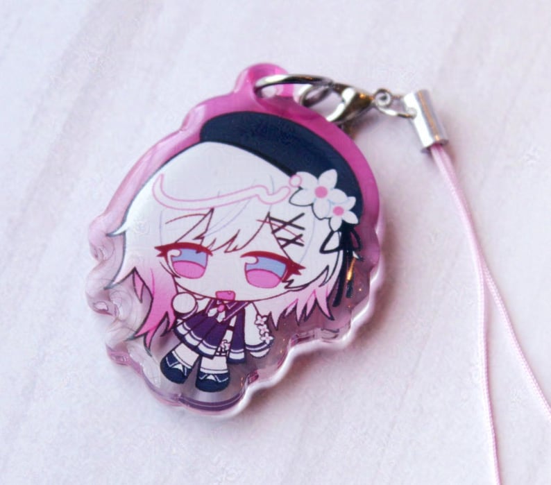 May include: A clear acrylic charm featuring a chibi-style character with white hair, pink highlights, and a black beret with a white flower. The character wears a dark purple outfit and is attached to a silver metal clasp with a light pink cord.