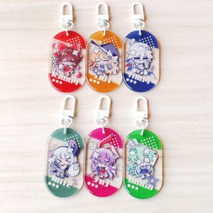 May include: Six oval acrylic keychains with white lobster clasps, each featuring a unique chibi anime character illustration. The keychains are colored red, orange, blue, teal, pink, and green, with printed text and three white stars at the base.