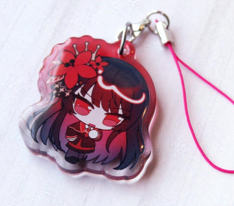May include: A clear acrylic charm featuring a chibi-style character with long dark hair, red eyes, and a red flower hair accessory. The character wears a dark red and black outfit. A silver metal clasp and a thin pink cord are attached to the top.