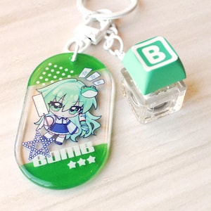 May include: A keychain featuring an oval green acrylic charm with a cartoon character illustration, a small frog, a blue star, and the text BOMB. Attached is a secondary charm consisting of a clear mechanical keyboard switch topped with a green B keycap.