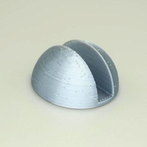 May include: A silver-colored, 3D printed, half-dome shaped phone stand with a slot for holding a phone.