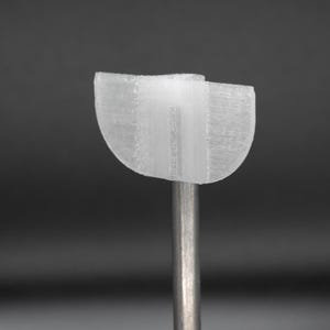 Może przedstawiać: Przezroczysty, półokrągły obiekt z pionowym wspornikiem. Obiekt wydaje się być drukowany w 3D, z widocznymi liniami warstw. Wspornik to cienki, srebrny pręt. Tło jest jednolicie ciemnoszare.