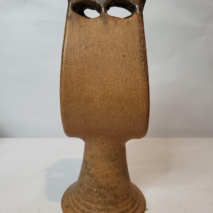 May include: A tall, brown ceramic vase with a unique, abstract design. The vase features a rectangular body with two eye-shaped openings near the top. The base is round and flared, and the overall height is approximately 12 inches.