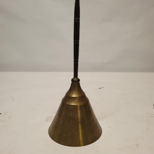 Pode incluir: Um sino de latão com uma alça de madeira escura. O sino tem uma forma cónica e um acabamento polido. A alça é longa e fina, com um topo arredondado. O sino é provavelmente usado para sinalização ou decoração.