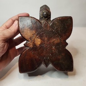 Puede incluir: Objeto de madera marrón oscuro con forma de mariposa. Presenta un diseño floral tallado en el centro y una pequeña decoración metálica. La veta de la madera es visible. El objeto parece hecho a mano. Podría ser un elemento decorativo.