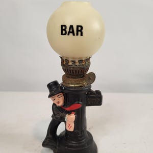 May include: A decorative lamp featuring a white spherical glass shade with the word BAR printed in black. The base is a black cast metal figure of a person in a top hat leaning against a post labeled BOURBON STREET, holding a small bottle.