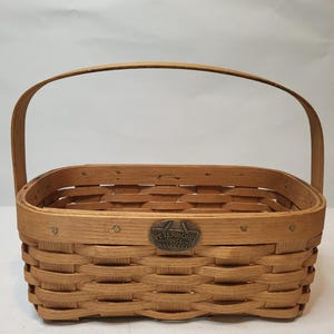 Könnte beinhalten: Ein rechteckiger Weidenkorb mit gebogenem Griff. Der Korb besteht aus hellbraunem Holz und hat ein Metallabzeichen mit der Aufschrift "Peterboro Basket Co.". Der Korb ist mit einem gewebten Muster gestaltet und eignet sich zur Aufbewahrung oder Präsentation.