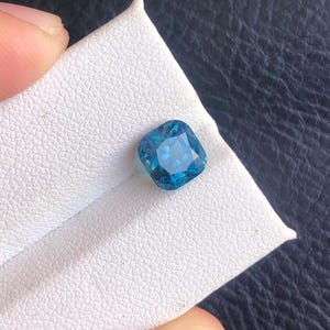 May include: A square-shaped, blue gemstone with a faceted cut. The gemstone is set on a white background.