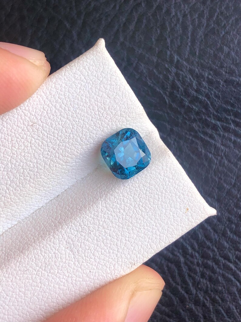 May include: A square-shaped, blue gemstone with a faceted cut. The gemstone is set on a white background.