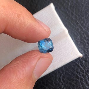 May include: A cushion-cut blue gemstone with a dark blue hue.