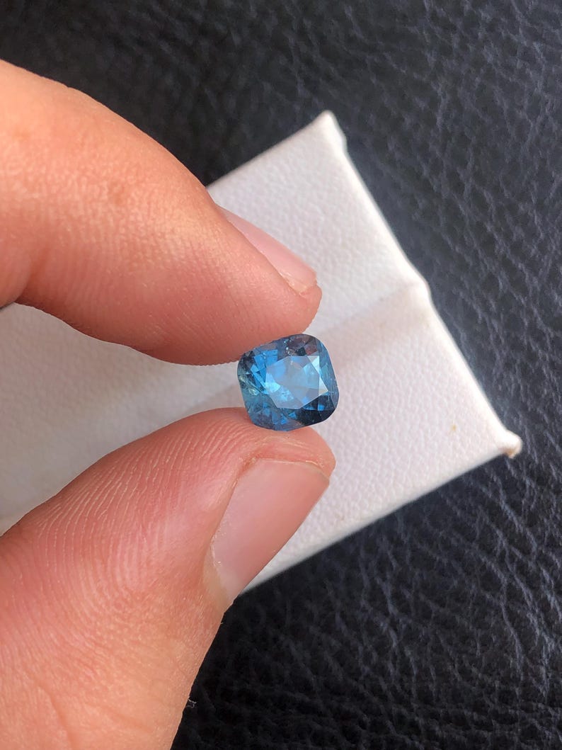May include: A cushion-cut blue gemstone with a dark blue hue.