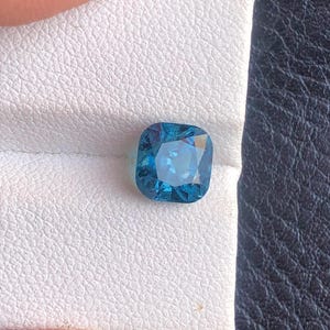 May include: A square-shaped, cushion-cut gemstone with a deep blue color. The gemstone is set on a white background.