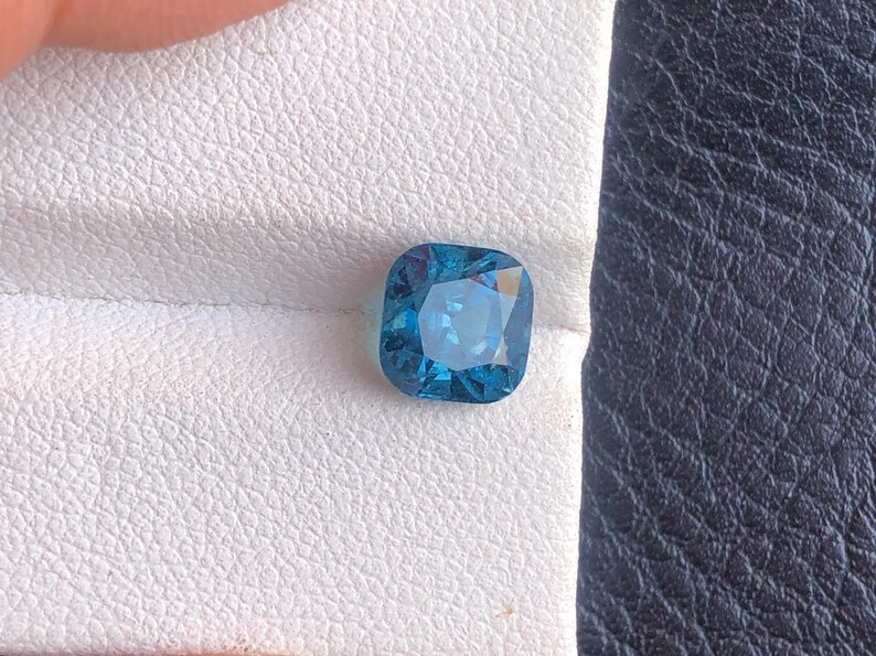 May include: A square-shaped, cushion-cut gemstone with a deep blue color. The gemstone is set on a white background.