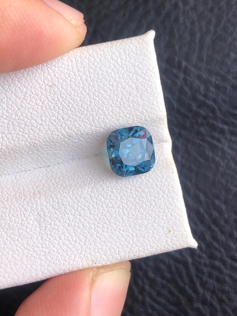 May include: A cushion-cut blue gemstone on a white background.