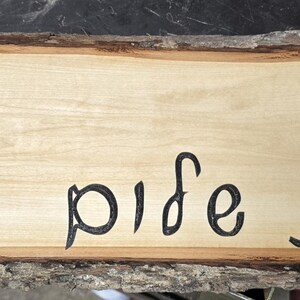 May include: A rectangular wooden sign with live edges. The sign is made of light-colored wood with dark brown edges. The Hebrew words "Shabbat Shalom" are carved into the wood in black lettering. The sign is approximately 24 inches long.