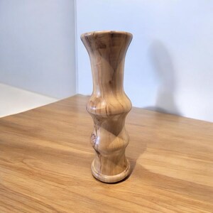 May include: A handcrafted wooden vase with a light brown finish. The vase has a unique hourglass shape with a flared top and base. The wood grain is visible, adding to its natural aesthetic. A decorative item for the home.