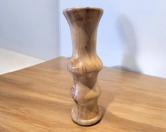 May include: A handcrafted wooden vase with a light brown finish. The vase has a unique hourglass shape with a flared top and base. The wood grain is visible, adding to its natural aesthetic. A decorative item for the home.