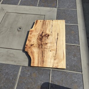 May include: A light-colored wooden cutting board with a unique, irregular shape. The board has a natural wood grain pattern with dark brown streaks and knots. It is placed on a gray tiled surface.