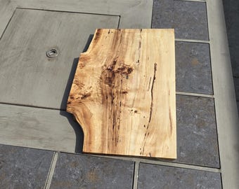 May include: A light-colored wooden cutting board with a unique, irregular shape. The board has a natural wood grain pattern with dark brown streaks and knots. It is placed on a gray tiled surface.