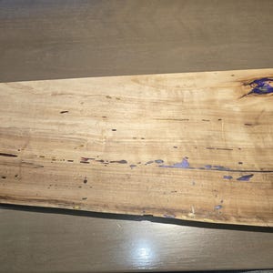 May include: A rectangular wooden serving board with a natural wood grain pattern. The board features dark brown edges and accents of purple resin. The wood has a light brown color with dark brown and purple accents.
