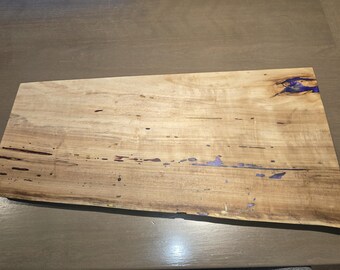 May include: A rectangular wooden serving board with a natural wood grain pattern. The board has dark brown edges and purple resin accents. The wood is light brown with dark brown and purple accents.