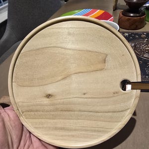 May include: A round, light-colored wooden serving tray with a raised edge. The tray has a natural wood grain pattern and a small, rectangular cutout. The tray is held by a person's hand.