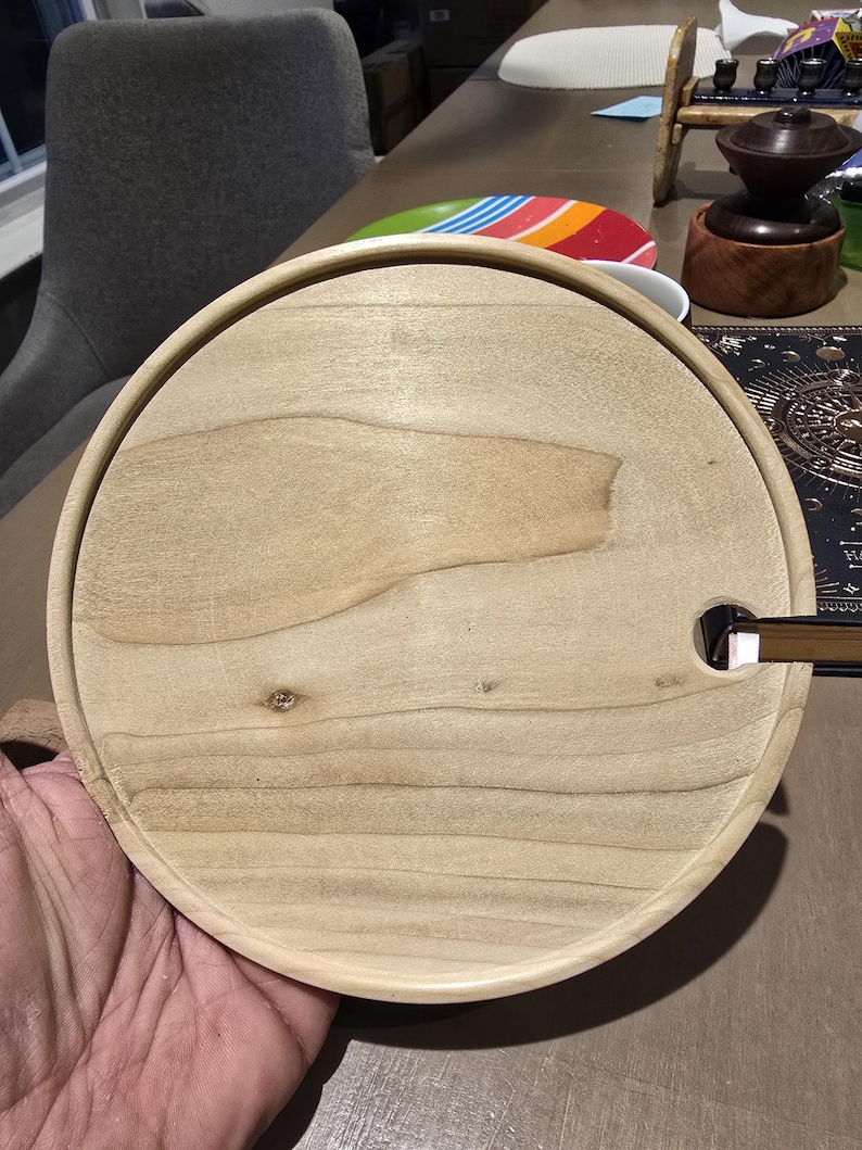 May include: A round, light-colored wooden serving tray with a raised edge. The tray has a natural wood grain pattern and a small, rectangular cutout. The tray is held by a person's hand.