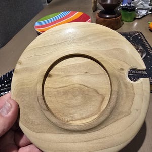 May include: A light-colored wooden coaster with a circular indentation in the center. The coaster has a smooth, polished finish and a small notch on the side. The wood grain is visible. The coaster is likely handmade.