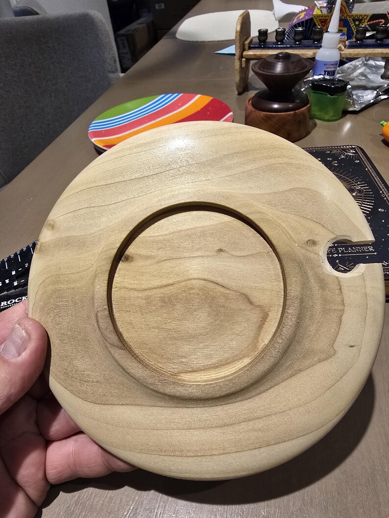 May include: A light-colored wooden coaster with a circular indentation in the center. The coaster has a smooth, polished finish and a small notch on the side. The wood grain is visible. The coaster is likely handmade.
