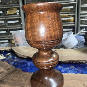 May include: A handcrafted wooden goblet with a dark brown finish. The goblet features a wide, flared top, a rounded midsection, and a sturdy base. The wood grain is visible, adding to the goblet's rustic charm. The goblet is likely used for drinking.