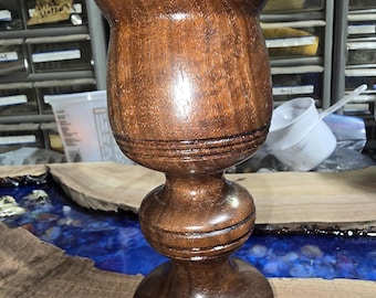 May include: A handcrafted wooden goblet with a dark brown finish. The goblet features a wide, flared top, a rounded midsection, and a sturdy base. The wood grain is visible, adding to the goblet's rustic charm. The goblet is likely used for drinking.