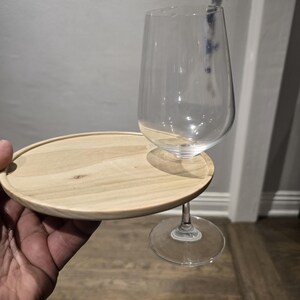 May include: A clear wine glass with a stem, resting on a small, round, light-colored wooden tray. The tray is attached to the glass stem, creating a portable wine glass holder.