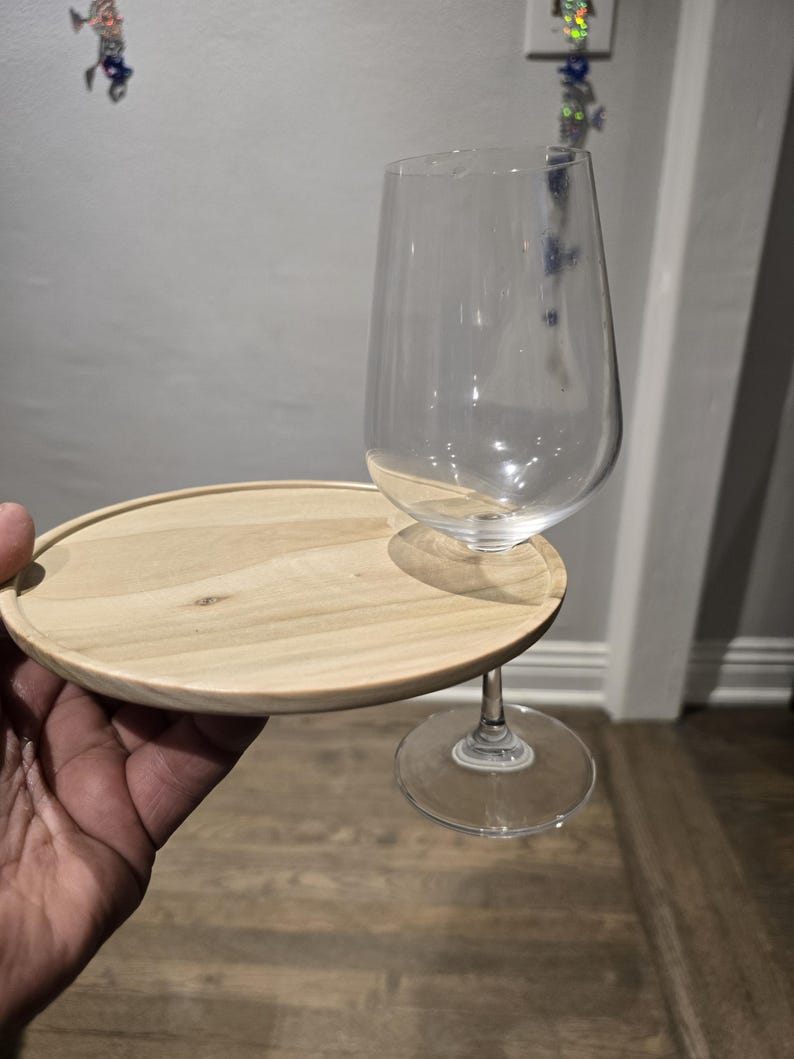 May include: A clear wine glass with a stem, resting on a small, round, light-colored wooden tray. The tray is attached to the glass stem, creating a portable wine glass holder.