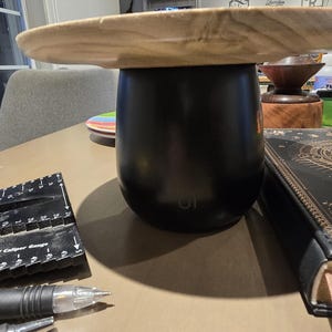 May include: A black tumbler with a wooden lid sits on a table. A black ruler and pen are in the foreground. A decorative book with gold accents and a small stack of colorful plates are also visible.