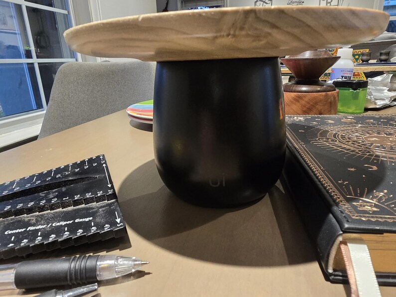 May include: A black tumbler with a wooden lid sits on a table. A black ruler and pen are in the foreground. A decorative book with gold accents and a small stack of colorful plates are also visible.