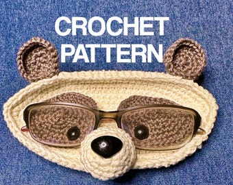 May include: Crocheted bear-shaped glasses holder with brown ears and a beige face. The holder has a pair of glasses resting on it. The text "CROCHET PATTERN" is written above the holder.