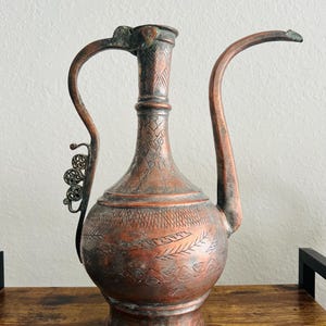 May include: An antique copper pitcher with an ornate design. The pitcher has a long, curved spout and a handle with a decorative metal piece. The pitcher is in good condition, with some signs of wear and tear.