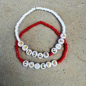 May include: Two beaded bracelets with the words "KANSAS CITY CHIEFS" spelled out in gold letters on white beads. The top bracelet is white and the bottom bracelet is red.