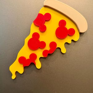 May include: A pizza slice magnet featuring a yellow melted cheese base, a tan crust at the top, and four red pepperoni toppings shaped like mouse heads. The design is flat with raised elements, showcasing a playful food-themed aesthetic.