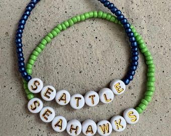 May include: Two beaded bracelets. One is navy blue, the other is green. The top bracelet spells out "SEATTLE" in white beads with gold lettering. The bottom bracelet spells out "SEAHAWKS" in white beads with gold lettering.